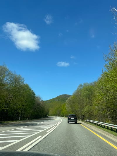 The image depicts a scenic road trip through a lush, mountainous landscape in Khrebtovoe, Russia, under a clear blue sky. The photo is taken from the perspective of a vehicle, looking forward on a paved, winding road.
The road features a solid yellow line marking the separation between lanes or the shoulder, and prominent white dashed lines on the right, indicating either a shoulder or an area for parking. A sturdy metal guardrail runs along the right edge of the road, protecting against what appears to be a drop-off or steep embankment. Ahead, a dark-colored car, possibly an SUV, is visible on the right lane, moving away from the viewer.
Both sides of the road are densely lined with vibrant green trees, their foliage suggesting either spring or summer. In the background, a large hill or mountain, also covered in rich greenery, rises against the horizon. The sky is a brilliant, unblemished blue, with a few sparse, white cumulus clouds scattered across it. The bright, even lighting indicates it is daytime, likely morning or afternoon, with excellent weather conditions for travel. The scene conveys a sense of peaceful journey through nature. There is no visible text in the image.