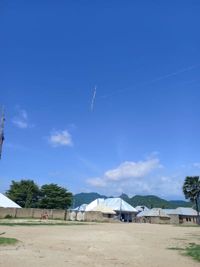 A wide shot captures a rural Nigerian village scene under a clear, bright blue sky with scattered white clouds. In the foreground, a dirt ground is visible with some patchy green grass. A wooden pole stands on the left, with what appear to be electrical wires stretching across the frame.
In the middle ground, a compound wall made of concrete separates a dirt area from a collection of buildings. Two individuals, possibly children, are seen near the wall, dressed in light-colored clothing. Beyond the wall, several structures with corrugated metal roofs are visible, some appearing to be homes. Trees, including two large, dense green trees, are situated behind the wall.
Further back, the landscape rises into rolling hills covered in greenery, suggesting a mountainous or hilly region. A palm tree stands tall on the right side of the image, adding to the tropical feel. The sun appears to be high in the sky, casting bright light and creating a daytime scene. The overall atmosphere is one of a peaceful, everyday rural life in Barakin Babele, Nigeria. No text is visible in the image.