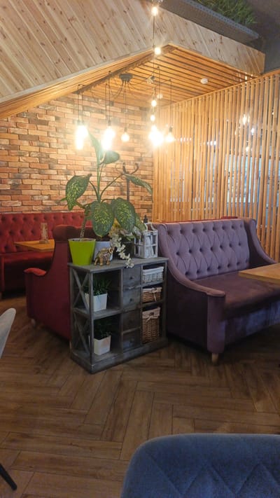 This media file captures an indoor scene, likely a cafe or restaurant in Langepas, Russia. The setting features a rustic and cozy ambiance, characterized by exposed brick walls, wooden paneling on the ceiling, and a herringbone-patterned wooden floor. Several plush seating areas are visible, including a deep red tufted banquette on the left and a purple tufted sofa on the right. Between the seating areas stands a distressed grey wooden shelf unit, decorated with a potted plant, a small decorative elephant, a white lantern, and woven baskets. The lighting comes from a cluster of Edison-style bulbs hanging from the wooden ceiling, suggesting it could be evening or a dimly lit interior. There are no people visible in the frame, and the overall mood is serene and inviting. The brick wall and wooden elements suggest a contemporary design with a touch of industrial or country style.