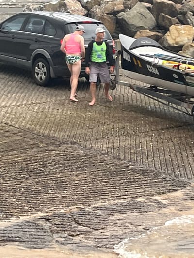 A dark gray vehicle backs down a concrete boat ramp toward the water. A boat on a trailer is hitched to the vehicle. Two barefoot individuals stand near the vehicle; one is wearing a bright pink top with patterned bottoms, and the other wears a bright green shirt and gray shorts. Large rocks are visible to the left, and the water's edge is to the right.