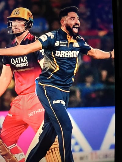 This is a photograph of a cricket match, likely taken during an Indian Premier League game given the team uniforms. In the foreground, two male cricket players are visible. The player on the right, wearing a dark blue and yellow jersey with "DREAM11" and "Raymond" logos, is in the process of bowling or celebrating a wicket, with his mouth open in a shout and his arms outstretched. He has a beard and a distinctive haircut. The player on the left is wearing a red and black jersey with "QATAR AIRWAYS" written on it and a yellow helmet with a protective cage. He appears to be watching the action. The background is blurred, showing spectators in the stands and a bright, possibly illuminated, stadium environment. There are no visible landmarks or features that would place this scene in Marusino, Russia.