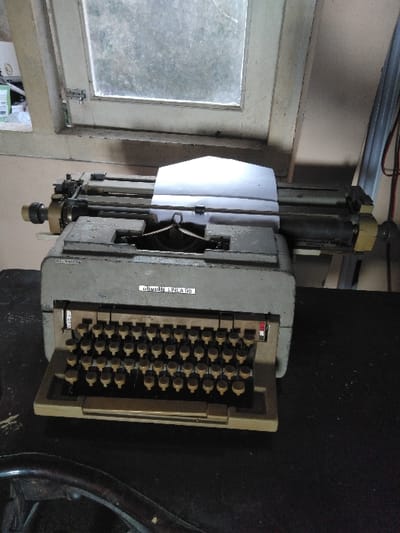 The scene is an interior view from Жібек Жолы, Kazakhstan. Dominating the foreground and mid-ground is a grey Olivetti Linea 98 manual typewriter, marked with its model name on the top cover. The typewriter features olive-green circular keys and has a sheet of plain white paper inserted into its platen. It rests on a dark, textured surface, with a curved, dark, possibly wooden object partially visible in the lower-left corner. Directly behind the typewriter is a two-pane window with a light-colored, worn wooden frame. The window panes are opaque or heavily obscured, preventing external visibility. A metallic latch is affixed to the left window frame. To the left of the window, a light-toned wall is visible, supporting several indistinct white containers on a narrow ledge. The far right background reveals dark electrical cables hanging vertically. No individuals are present.