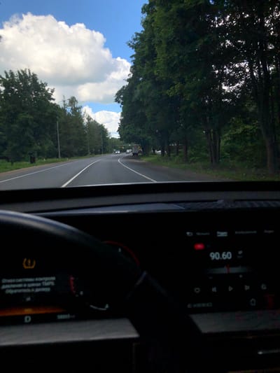 The image shows a view from inside a vehicle, looking out at a rural road lined with trees. The road is asphalt and has a white line marking the lanes. There are a few vehicles visible in the distance on the road. The sky is blue with large white clouds. The scene appears to be set in a rural area, possibly in Russia, given the location context. The time of day appears to be daytime, with good lighting and clear weather. The perspective suggests the photographer is a passenger or driver in the car.
On the right side of the image, part of the car's interior is visible, including a dashboard with a digital display. The display shows the time "09:06" and some other icons and text that are difficult to read clearly due to the image quality and angle. There is a red indicator light illuminated on the dashboard. Another part of the dashboard shows a circular dial and some text in Russian, which is also blurry. The overall impression is a snapshot taken during a drive through a scenic, tree-lined road on a sunny day.