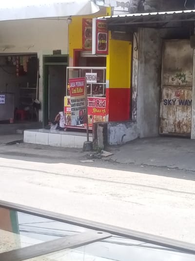 The image shows a street scene in Kecamatan Waru, Indonesia, located at latitude -7.35 and longitude 112.764. A person is sitting on the ground, leaning against a wall, in front of a small, red, yellow, and green storefront. The storefront is labeled "Jasa Permak Umi Ridho" and displays a sign for "Sorean". The building next to the storefront has a large, rusted metal door labeled "Sky Way". The street is made of concrete, and the ground is dusty. There is a gray, concrete sidewalk leading up to the storefront. The person sitting on the ground is facing the storefront. The sidewalk is composed of white square tiles. A small, green plant is growing on the side of the building next to the storefront. The image is taken from a low angle, looking across the street. The storefront is decorated with a colorful awning and several signs. The image is somewhat blurry.