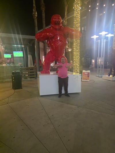 A middle-aged man in a pink t-shirt and black pants stands in front of a large, bright red gorilla sculpture at night in Burbank, California. The sculpture is on a white pedestal. To the left of the sculpture, there is a black trash can, some palm trees, and two televisions displaying a soccer game. To the right of the sculpture, there are string lights, street lamps, and a building with windows. A sign on an easel to the right of the sculpture reads "PARKLET RULES". The ground is paved with concrete.