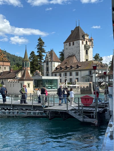The image depicts the pier in front of Oberhofen Castle, located in Oberhofen am Thunersee, Switzerland. Several individuals are standing on the wooden pier, some appear to be tourists. The architecture of the castle is visible in the background, characterized by its distinctive towers and pitched roofs. The water in the foreground is a deep blue, suggesting a lake or river. The sky is partly cloudy with a bright blue hue. The overall scene captures a sunny day at a scenic historical site.