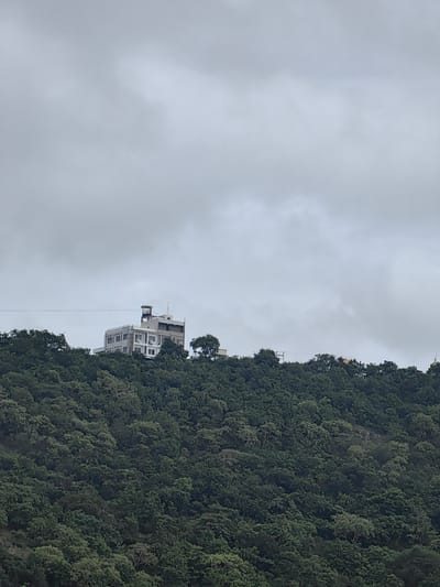 A building with a tower is perched on a heavily wooded hill under a cloudy sky. The building is white with numerous windows and appears to be several stories tall. A dark cable runs horizontally across the frame just above the building and the trees. The hill is densely covered with green trees, obscuring much of the terrain. The sky is overcast with gray clouds. The image captures an elevated view of the building and the natural landscape surrounding it.