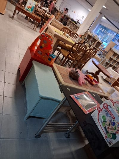A tilted, slightly high-angle shot captures an indoor scene with a variety of objects. Several wooden tables and chairs occupy the space. A colorful children's play structure is visible. A black table with chrome legs hosts a toy box and a framed photo. The background reveals shelves stocked with items in a retail-like setting, under bright, natural light. The overall impression is a busy, somewhat cluttered interior.