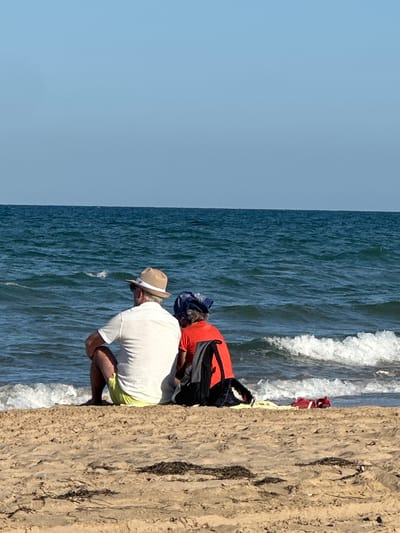 The image depicts two individuals, viewed from behind, seated on a sandy beach facing a calm, blue ocean under a clear sky. The person on the left wears a white polo shirt, light yellow shorts, and a straw hat. The person on the right wears an orange t-shirt and a blue bandana. A backpack rests between them, and a red item, possibly a towel or blanket, is on the sand to their right. The scene appears to be a tranquil moment at the beach. The location is Guardamar del Segura, Spain, a coastal town known for its extensive beaches and pine forests.