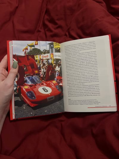 An open book displays a photograph of a red Ferrari 512 S race car with its hood and engine cover open, parked on a track. The car has the number 6 in a white circle on its side. In the background, there are spectators, flags, and grandstands with various sponsor logos, including "Shell." The book's text on the right page is in Russian, discussing the car's history and a race. There is no indication of the city of Vysokaya Gora, Russia in the image or text.