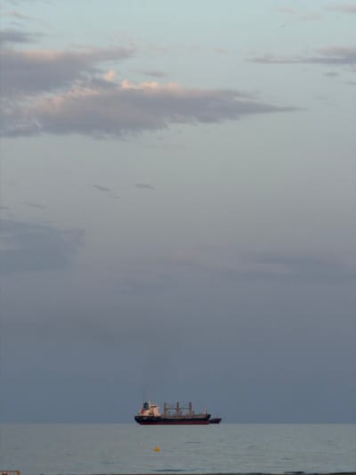 The image shows a cargo ship sailing on a calm sea. The sky is overcast with soft, diffused light, and there are a few scattered clouds. The sea is a muted blue-gray, reflecting the sky. The ship is in the distance, and its details are somewhat indistinct due to the lighting conditions and distance. A small yellow buoy is visible in the water closer to the foreground. The foreground itself is not clearly visible but appears to be a beach or shoreline. The scene suggests a tranquil, possibly early morning or late evening atmosphere, typical of coastal areas. The location is specified as Grao, Spain.