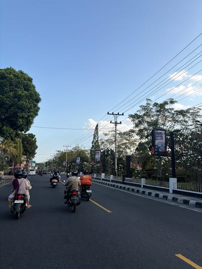In Magelang, Indonesia, on a clear day with a blue sky and scattered clouds, several people are riding motorcycles on a paved road. The road has yellow lane markings and a black and white striped curb. On the left side of the road, there are trees and buildings, including a sign for "Aroma" and another for "Food." On the right side, there are more trees and a white fence. The motorcycles are moving away from the camera, and some of the riders are wearing helmets. One motorcycle has a license plate that reads "K 6099 APC." Another has a license plate that reads "AA 3328 MC." The scene appears to be a street in a city or town.