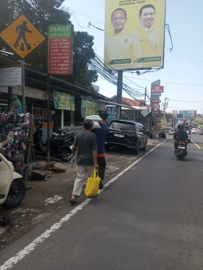 The image shows a street scene in an urban environment with several individuals, vehicles, and signs. In the foreground, two people are walking along the edge of the road, with one carrying a yellow shopping bag. A pedestrian crossing sign is on the left, and a restaurant sign for "Family Soto Ibu Imas" lists various Indonesian dishes and juices. A black car is parked on the side of the road, and motorcycles are parked nearby. A political billboard displays the images of two Indonesian politicians. Further down the street, there's moderate traffic with more motorcycles and cars, along with various storefronts and commercial signs. The sky is clear, suggesting it's daytime. The scene appears to be taken from a slightly elevated position.