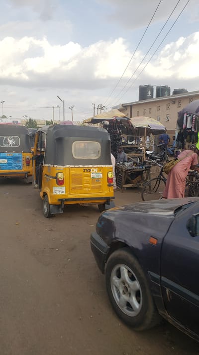 The image depicts a street scene in Kawo, Nigeria. A yellow-and-black tricycle, commonly known as a "Keke Napep", is parked in the street. To the right, the front quarter of a dark blue car is visible. In the background, there are shops and stalls selling various items, with people milling about, including one person on a bicycle and dressed in light pink robes. The sky is overcast with some visible clouds, and the road surface appears to be paved. Three black water tanks can be seen on top of a building. Overhead power lines run across the top of the frame.
