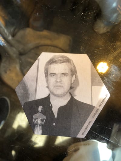 A close-up shot shows a hexagonal coaster lying on a dark, reflective surface. The coaster features a black and white image of a man in a buttoned shirt holding what appears to be an Oscar statue. Printed along the edge of the coaster is the text "HR GIGER BAR - GRUYERES - SWITZERLAND." The overall tone of the image is subdued due to the black and white aesthetic and the dark background. The surface beneath the coaster shows signs of wear and reflections of light sources. The bar is in Gruyeres, Switzerland. Epagny is nearby.