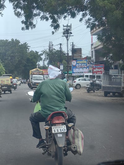 In the city of Datala, India, on a cloudy day, a man wearing a green shirt and black pants rides a motorcycle on a paved road. He has a white cloth covering the back of his head. A striped bag is hanging from the rear of the motorcycle. The road is busy with traffic, including trucks, cars, and other motorcycles. Several buildings and trees line the street, and electrical wires are visible overhead. Billboards advertise various services, including education and medicine.