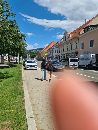 A medium shot captures a street scene in Sankt Marein bei Neumarkt, Austria, on a clear day with scattered clouds. Two young women walk away from the camera along a brick sidewalk. The woman in front wears a black t-shirt and black shorts, with her dark hair in a braid. The woman behind her wears a black t-shirt and light blue jeans, with her dark hair in braids. They are walking on a sidewalk bordered by a grassy area on the left. Across the street, a row of buildings with colorful facades line the road. Several vehicles are present on the street, including a white Skoda, a dark gray hatchback, two vans, and a truck. A white van with "Glaserei Zischka" written on it is parked on the right side of the street. In the background, a green hill with scattered trees and houses is visible under a bright blue sky. The overall impression is a sunny day in a European town.