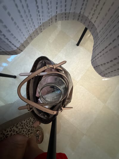 A high-angle, eye-level shot shows a brown leather tote bag resting on a light-colored tiled floor. The bag contains a clear glass jar and some light-colored items, possibly clothing. A person's legs and feet are visible in the lower left of the frame, one leg clad in a leopard-print material and the other in a red garment. The person is wearing a sandal adorned with cartoon characters. The scene appears to be indoors, possibly under a table or desk, as a patterned, sheer curtain is draped from above, casting shadows. The floor tiles are arranged in a grid pattern. The lighting is dim, with some areas brighter than others due to artificial light sources. There are no identifiable landmarks or features to indicate a specific location in Rome, United States of America.