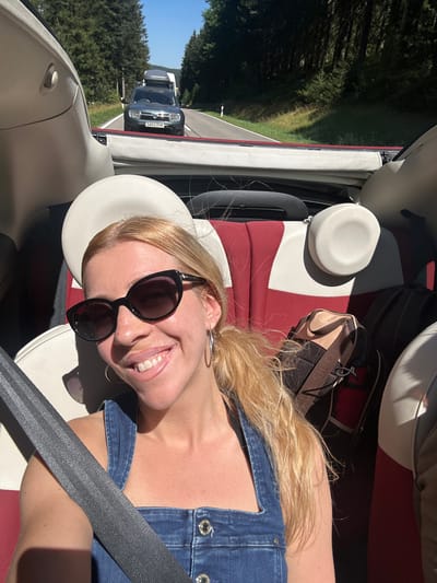 In the city of Furtwangen im Schwarzwald, Germany, a woman with blonde hair, wearing sunglasses and a denim jumpsuit, is taking a selfie in the passenger seat of a convertible. She is smiling and looking at the camera. The car is driving on a road through a forest. In front of the car, there are other vehicles on the road. The seats of the convertible are red and white. There are also backpacks in the car. The sky is blue and the sun is shining.