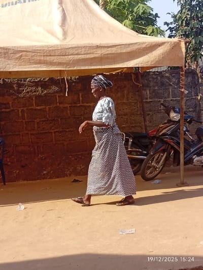 An elderly woman wearing a headscarf, light-colored blouse, and checkered skirt walks along a sandy path. She wears sandals and her right arm is bent. A tan awning covers her, attached to wooden posts. Behind her, a low brick wall and a motorcycle are visible. Litter is scattered on the ground. The date "19/12/2025 15:24" is printed in the lower right corner.