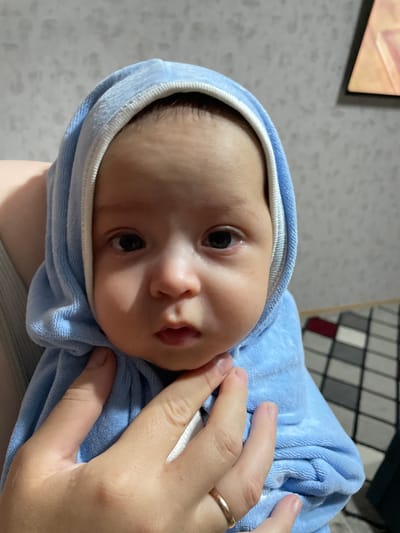 A close-up shot features a baby swaddled in a light blue hooded towel. The baby's face, with dark eyes, a small nose, and slightly parted lips, is the central focus. A person's hand, wearing a ring on the left ring finger, is gently supporting the baby's chin and neck, suggesting an interaction of care. The background is slightly blurred, showing a wall with a textured pattern and a portion of what appears to be a rug with a checkered design. There is also a framed picture on the wall in the upper right corner of the image. The lighting appears to be indoor, creating soft shadows on the baby's face. The scene does not provide any specific visual cues that would allow for the identification of the location within the city of Fălești, Moldova.