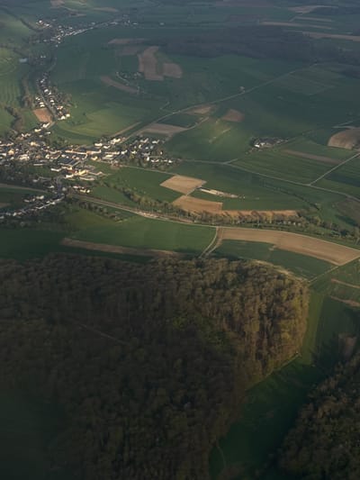 The image depicts an aerial view of the city of Berg, Luxembourg, characterized by a rural landscape with a mix of green fields, forested areas, and a dense urban area. The fields exhibit varying shades of green, suggesting different crops or stages of growth. A dense forest borders the fields. The town is densely packed with buildings. A network of roads and paths weaves through the landscape, connecting the different areas. The lighting suggests the image was taken during daylight hours.