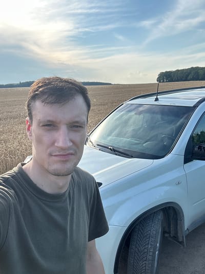 A man in a green t-shirt takes a selfie outdoors in a field of wheat. He is standing next to a white SUV. The sky is partly cloudy. The location is the city of Slobidka-Zalisetska, Ukraine.