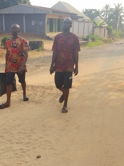 Two men walk on a sandy road. The man on the left wears a red and gold patterned shirt, black shorts, and sandals, carrying a black bag. The man on the right wears a red and black patterned shirt, black shorts with white letters on the leg, and sandals. A building with a gray roof and white walls is in the background. Palm trees are visible in the distance, on the right.