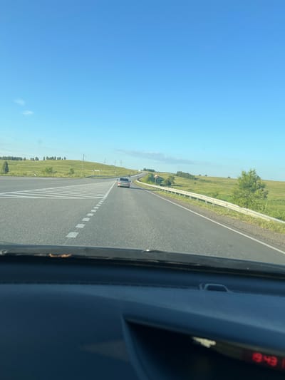 The image captures a view from inside a car driving on a paved road in Kopalovskii, Russia. The road curves to the right and is bordered by a metal guardrail on the right and a grassy field on the left. There are green hills in the distance, dotted with trees and interspersed with patches of sky. Several power poles are visible along the road. A silver car is driving ahead in the same direction. The sky is clear and blue, with a few wispy clouds. The dashboard of the car is partially visible in the foreground, with a digital display showing "19:43".