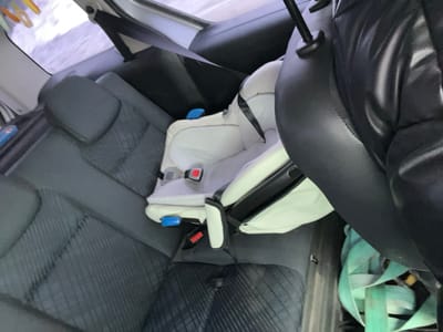 The scene depicts the interior of a vehicle, specifically the rear passenger compartment, located in the city of Podvyaznovo, Russia. A light-colored, unoccupied infant car seat is secured on the rear passenger seat directly behind the front passenger seat. The rear seats feature dark grey fabric upholstery with a quilted or textured pattern. The back of the front passenger seat, upholstered in black material, is partially visible on the right. In the immediate foreground, a segment of a person's upper body or arm, wearing a black puffy jacket, is visible. On the vehicle floor in the foreground, a bundle of light blue or teal webbing straps is present. Through the rear window, a blurry, bright, light-toned exterior environment is visible, consistent with an overcast sky or snowy conditions. No individuals are depicted occupying the car seat or directly interacting within the visible frame.