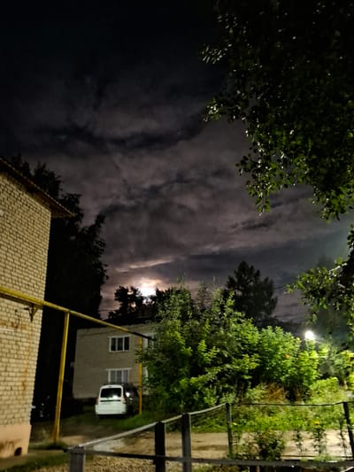 This is a night-time photograph of an urban residential area, likely in Novlyanka, Russia, with a cloudy sky obscuring most of the moon. In the foreground, a brick building is partially visible on the left, with a yellow pipe running horizontally. To the right of this building, a smaller, lighter-colored building with two windows on the upper floor is visible. A white van is parked in front of this building. Dense green foliage, including trees and bushes, dominates the middle and right side of the image. A street lamp is illuminated among the trees on the right. The ground appears to be wet, possibly indicating recent rain. A metal railing runs across the bottom of the frame.