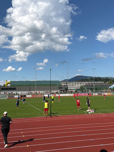 The image shows a sports field with a running track, located in Rapperswil-Jona, Switzerland, based on the presence of Swiss flags and local advertisements. Several individuals wearing soccer uniforms in red and blue colors are on the field. A person dressed in black attire and a white cap is positioned on the track, casting a shadow. The background includes buildings, a grassy area, and a clear sky with clouds. There are light poles lining the field. The scene suggests an athletic event or training session taking place.