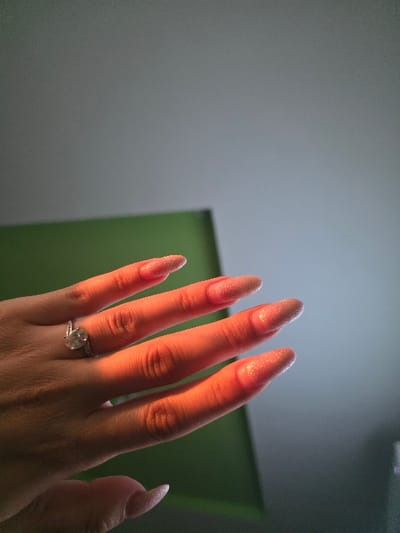 An image taken in Staliyska mahala, Bulgaria, depicts the dorsal aspect of a left human hand. The hand exhibits extended, almond-shaped artificial nails with a light, shimmery, possibly pink or nude glitter finish. A silver-toned ring with a prominent oval or cushion-cut central stone, flanked by smaller stones, is worn on the fourth digit. Strong, warm-toned light illuminates the hand from the left, creating an orange hue on the skin and distinct shadows between the fingers. The background is out of focus, featuring a large, flat, dark green surface to the lower-left and center, and a solid light grey surface above and to the right. No other individuals or actions are visible.