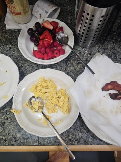The image depicts a breakfast scene on a speckled granite countertop. In the foreground, a white plate holds a serving of scrambled eggs, with a spoon resting inside. To the right of this plate, another white plate is covered with a paper towel, on which two strips of cooked bacon are placed. In the background, a white bowl overflows with fresh strawberries, raspberries, and cherries. Adjacent to this bowl, a metal utensil holder stands filled with various kitchen utensils. A glass bottle of marinara sauce and a partially unrolled paper label are also visible on the countertop. The overall environment suggests a domestic kitchen setting. There is no information in the image to indicate a specific location in Seattle, United States of America.