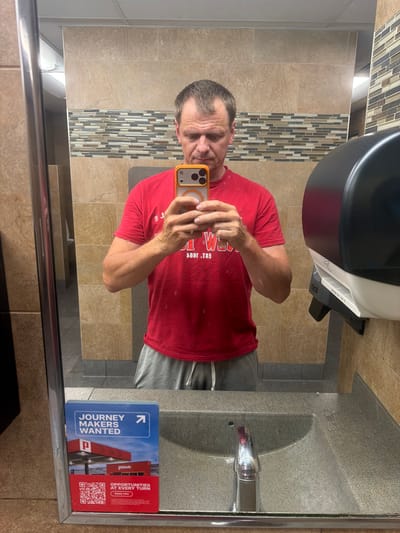 A man is taking a mirror selfie in a public restroom. He is wearing a red t-shirt and grey sweatpants, holding a smartphone in his right hand. In the reflection, the restroom features beige tiled walls, a patterned tiled accent wall, a sink with a chrome faucet, and a black and white paper towel dispenser. On the left side of the mirror, a "JOURNEY MAKERS WANTED" poster with a QR code and a Pilot gas station image is visible. The man's face is slightly obscured by his phone. The setting appears to be a truck stop or rest area, possibly in Lebanon, United States of America, given the context of the "Pilot" branding.