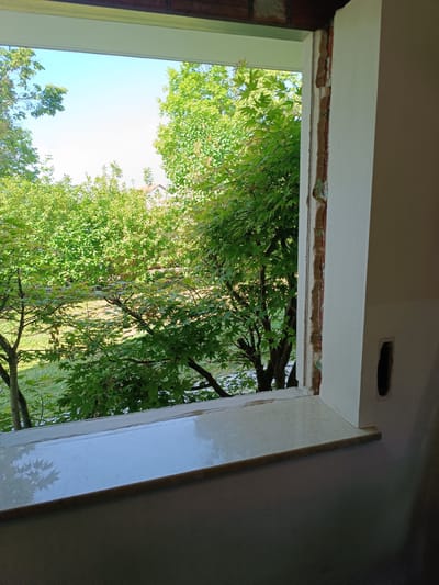 A view from inside a building, looking out of a window. The window frame is white with some unpainted areas visible on the right side. Outside, there are trees with green foliage, suggesting a natural or residential area. A building can be seen in the background, partially obscured by the trees. The window ledge appears to be made of tile. There are no people visible in the scene. The location is Essenbach, Germany.