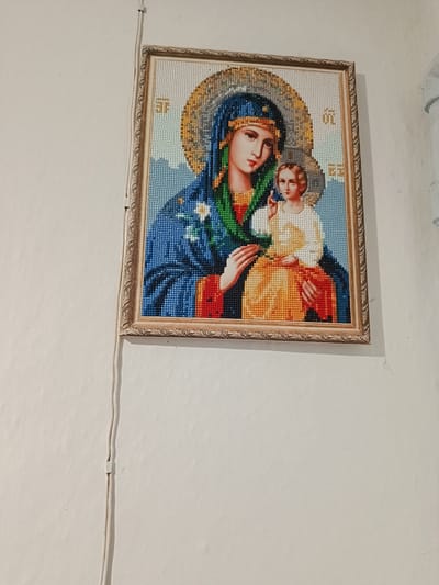 The image shows a framed religious icon, depicting the Virgin Mary holding the infant Jesus. The icon is a diamond painting, characterized by its mosaic-like appearance created by small, colored rhinestones. The Virgin Mary is dressed in a blue robe and a blue head covering, with a green sash. She is looking downwards at the infant Jesus, who is reaching out with one hand. The background of the icon is a mosaic of light blue and white beads, with a golden halo around the figures. The icon is hanging on a white, textured wall. A white electrical cord runs vertically down the wall next to the icon, attached by a clip. The location is specified as Койлык, Kazakhstan, suggesting this image was taken in a religious or residential setting within that city.
