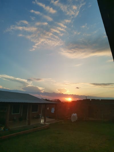 The image depicts a backyard scene at sunset in the city of Obligado, Paraguay. A single-story brick house with a metal roof is visible on the left, featuring a covered porch with pillars. In the background, a brick wall under construction stands to the right of the house. The yard is covered with green grass, and a few objects, including what appears to be a white tarp covering something, are scattered around. The sky is a vibrant mix of blue and orange hues, with scattered clouds and sun rays emanating from the setting sun, which is partially obscured by clouds. The overall impression is that of a residential area experiencing the end of a day.