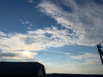 A dusk sky with scattered clouds over Sharan, Russia, is captured in this photograph. The image features a portion of a dark-tiled roof in the lower left corner, likely belonging to a residential building. A utility pole with wires is visible on the right side, extending towards the top of the frame. In the distance, a dark, silhouetted treeline marks the horizon. The sun is setting, casting a warm glow through the clouds, creating visible sunbeams. The overall environment appears to be a rural or suburban setting on the outskirts of the city. There are no people or animals visible in the frame.