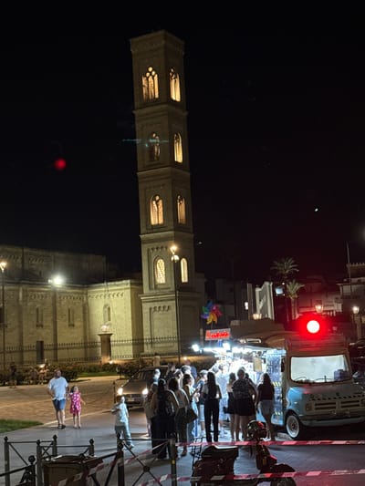 The image captures a nighttime scene in Parabita, Italy, featuring the illuminated bell tower of the Church of Sant'Antonio Abate. The tall, rectangular stone tower stands prominently, showcasing multiple levels of arched windows emitting a warm yellow light. To the left, a section of the church's stone facade is visible, less brightly lit, bordered by a black metal fence.

In the mid-ground, to the right of the tower, a light blue, vintage-style food truck is parked, distinguished by a bright red spherical lamp on its roof. Approximately 15-20 individuals, including adults and children, are gathered in a queue or close proximity around a brightly lit food stand situated directly to the left of the truck. This food stand features a canopy with a colorful pinwheel sign.

On the left side of the frame, a man in a white t-shirt and blue shorts walks with a child in a pink patterned dress. Another child in a light-colored shirt and pants stands closer to the crowd. The immediate foreground shows a paved area with metal barriers striped in red and white, behind which two dark-colored scooters or mopeds are parked. Streetlights provide additional illumination, and several palm trees are visible against the dark sky in the background. A dark car is partially visible behind the pedestrians.