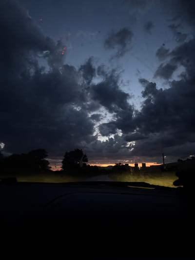 The image depicts a view from inside a vehicle looking out at a dramatic sky at dusk or twilight. Dark, heavy clouds dominate the upper portion of the frame, with shafts of light breaking through in the distance, suggesting a sunset or the last rays of daylight. The horizon shows silhouetted trees and what appear to be industrial structures, possibly cooling towers, emitting steam or smoke, illuminated by the orange glow of the setting sun. The foreground is very dark, obscuring the interior of the car. There is no information in the image that allows for identification of the location as Balanovo, Bulgaria.