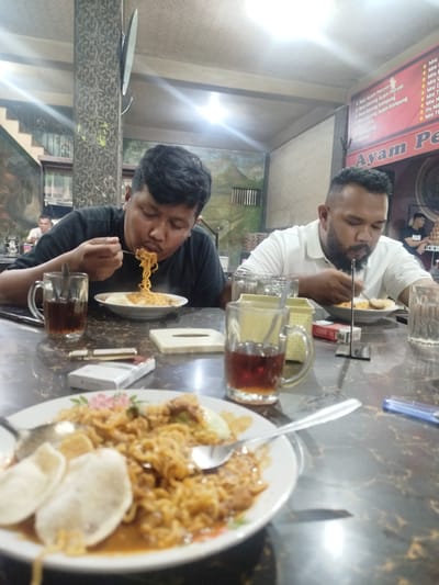 Two men are seated at a table in a restaurant, each eating a plate of noodles. The man on the left is wearing a black shirt, while the man on the right is wearing a white shirt. Both men are intently focused on their meals. The table is covered with various items, including cups of tea, cigarette packs, lighters, and a plate of noodles and crackers in the foreground.

The restaurant appears to be in the city of Kutalhoksukon, Indonesia, as indicated by the restaurant's sign "Ayam Pe" which is partially visible. A menu board lists local dishes such as "Nasi Ayam Penyet" and "Mie". The interior is dimly lit, with decorative wallpaper and artwork on the walls. Other customers can be seen in the background.