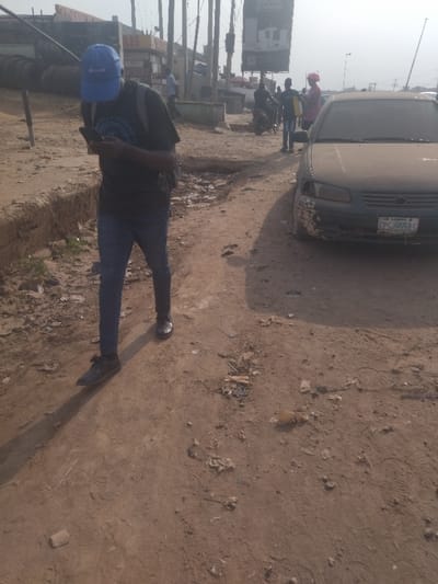A man wearing a blue hat and a black shirt is walking down a dirt road in Way, Nigeria, while looking at his phone. The man is walking towards a silver sedan parked on the right side of the road. Other people are walking on the sidewalk in the distance. The ground is dusty and covered in debris. The road is rough and uneven. There is a large billboard on the right side of the road. The sky is cloudy.