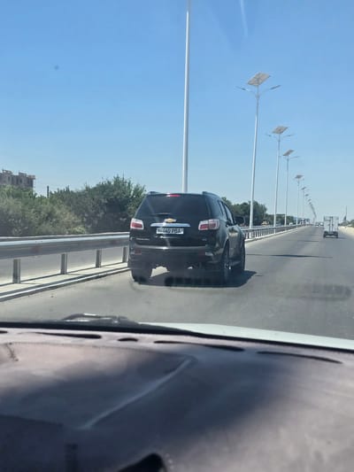 A black Chevrolet Trailblazer is seen driving on a highway on a clear, sunny day. The vehicle is in the foreground, with its license plate visible and reading "60 440 PBA". The highway is lined with metal guardrails and streetlights with solar panels. In the background, there are trees and buildings, including what appears to be an unfinished construction site. The perspective is from inside another vehicle, looking out through the windshield.
