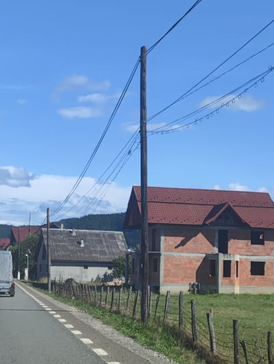 The image depicts a rural scene in Petrova, Romania, under a bright, partly cloudy blue sky, suggesting it is daytime and the weather is fair. The foreground shows a paved road with white dashed lines, bordered by a grassy verge and a rustic wooden fence.
Several houses are visible. To the left, a light-colored, older house with a grey, corrugated roof stands next to the road. Further down the road, a white truck is partially visible. To the right of the older house, a new, unfinished house constructed from red brick is prominent. It features a red tiled roof and has dark, empty window openings. Behind these structures, a dense line of evergreen trees forms a backdrop, indicating a forested area.
Overhead, a wooden utility pole supports multiple power lines, some of which are adorned with small, circular insulators. The overall impression is one of a developing rural community, with older dwellings alongside new construction. There is no visible text in the image.