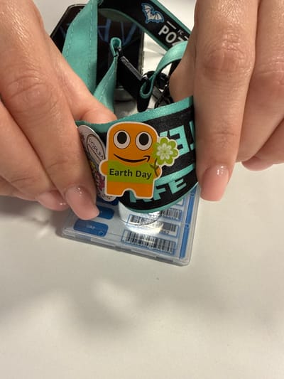 A close-up, eye-level shot shows two hands, likely belonging to the same person, interacting with a lanyard and ID badge. The skin tone suggests a light complexion. The nails are manicured with a subtle, pale pink polish, neatly shaped.
The lanyard is a vibrant teal with black accents. It features a black ribbon with white text and a graphic of a winged creature. Attached to the lanyard is a clear plastic ID card holder containing multiple items, including what appear to be barcodes or serial numbers printed on white strips.
Prominently displayed on the lanyard is a colorful sticker or charm. It's an orange, cartoonish character with large black eyes and a green flower with four petals on its head. The text "Earth Day" is visible in green on the character. A smaller circular sticker below it contains a logo with the word "FORUM" and other unreadable text.
The hands are shown gently holding and manipulating the lanyard and badge, suggesting someone is either attaching or adjusting it. The lighting is bright and even, indicating an indoor setting, likely during the daytime. The background is a plain, light-colored surface, possibly a desk or table, with subtle texture. The overall mood is neutral and practical, focusing on the details of the items. There are no cues about the weather or a specific time of day beyond "daytime."
The visible text includes "POZ" on the lanyard and "Earth Day" on the charm. The location context of Chociule, Poland, does not offer any direct visual cues within the image itself.