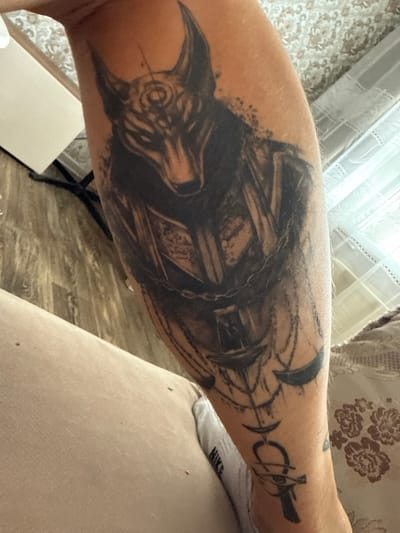 A close-up, eye-level, medium shot captures a tattoo on a person's forearm. The tattoo is a detailed, dark ink illustration of Anubis, the ancient Egyptian god of the afterlife, depicted in a realistic, stylized manner. Anubis's head is prominent, with sharp ears and intricate facial markings. The body of the tattoo features elements like chains and what appears to be a scale, suggesting themes of judgment or passage. The tattoo extends down the forearm, with the lower portion displaying an ankh symbol and the Eye of Horus. The skin tone of the arm is visible around the tattoo, and fine hairs can be observed. The background is blurred, but suggests an indoor setting with a textured surface that may be flooring or upholstery, and a white, possibly translucent object on the left. No specific details of Kondopoga, Russia are visible in this image.