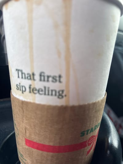 A close-up shows a Starbucks coffee cup with a cardboard sleeve. "That first sip feeling." is printed vertically on the cup. The sleeve, mostly brown, features a red ribbon design with green Starbucks lettering. Coffee stains mark the white cup near the printed text. Part of a dark-colored object, possibly a car interior, is visible at the bottom.