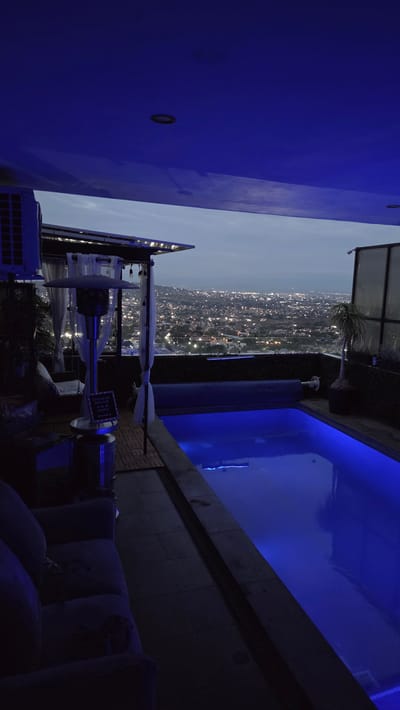 The scene captures an evening view in Tonalá, Mexico, featuring a rooftop setting with a pool illuminated by blue lighting. A partial cityscape is visible in the background, showcasing numerous lights, indicative of a dense urban area. Foreground objects include a partially covered patio heater, and bags of cement or other construction materials. A low wall separates the foreground from the pool area. The pool water is brightly lit, contrasting with the darker areas of the scene. A tall building wall, also lit in blue, dominates the left side of the image.