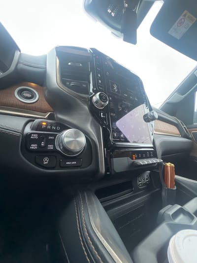 This media file captures a close-up, low-angle view of the interior of a modern vehicle, likely a truck or SUV, in Jõhvi, Estonia. The central focus is the dashboard, dominated by a large touchscreen infotainment system displaying navigation or media information. To the right of the screen, a rotary dial controls the four-wheel-drive system, with labels indicating modes such as "2WD," "4WD LOW," "4WD AUTO," and "4WD HIGH." Below this, a button labeled "AXLE LOCK" is visible.
Further to the right, a cup is placed in a holder. The dashboard also features wood grain trim, adding a touch of luxury. A prominent "START ENGINE STOP" button with a circular design is situated on the center console. Above the dashboard, a sun visor is flipped down, displaying an "AIRBAG" warning label. The overall setting appears to be daytime, with bright, overcast sky visible through the windshield, suggesting potentially cloudy weather. No people are visible in the frame, emphasizing the focus on the vehicle's internal features and controls. The image is sharp and well-lit, highlighting the details of the car's interior design and technology.